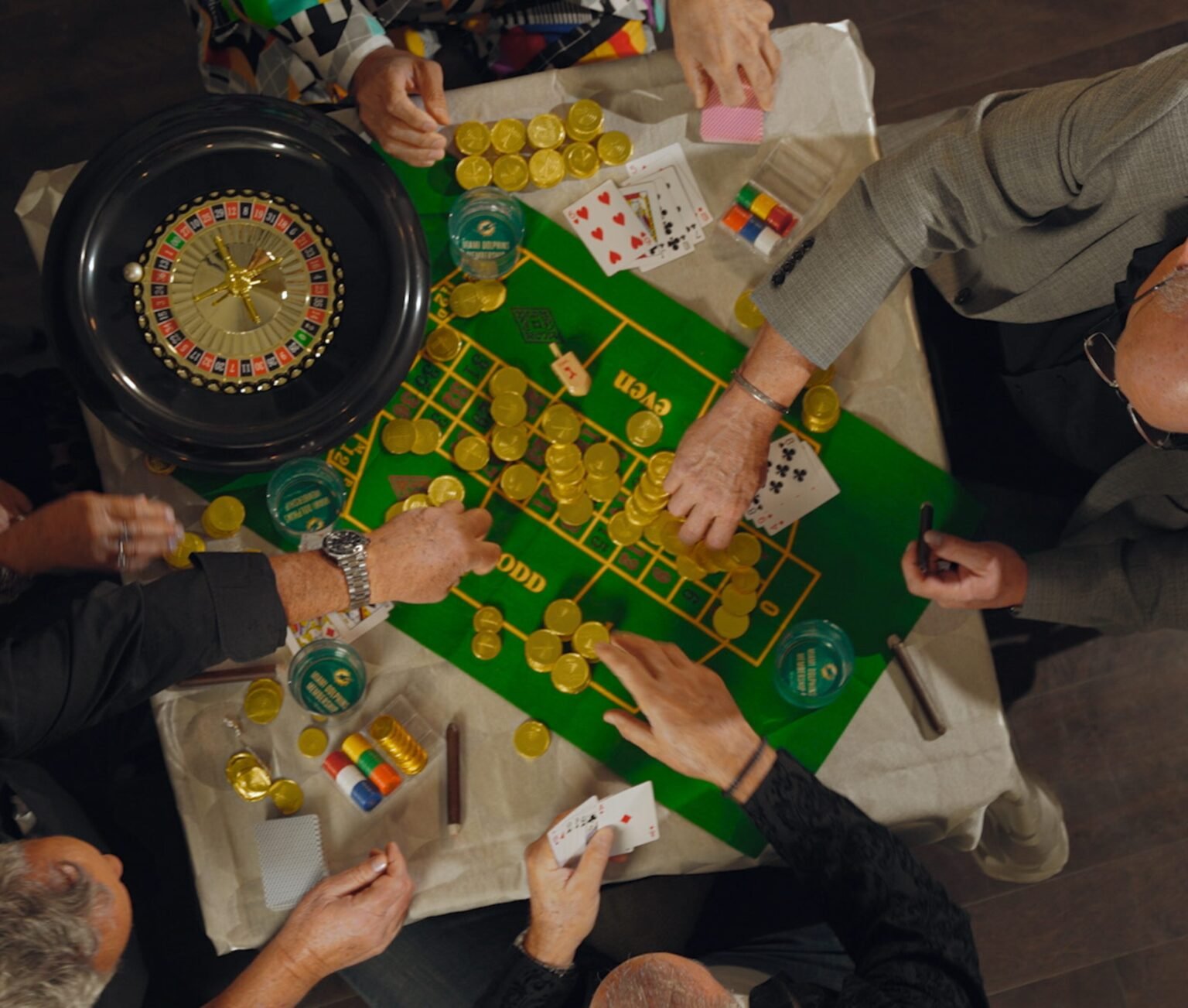 Overhead view of hands around a green roulette and craps layout piled with gold chips